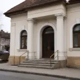 O clădire cu arhitectură administrativă clasică, amintind de o primărie, cu coloane la intrare, uși duble din lemn și ferestre boltite