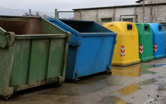 O serie de containere de colectare a deșeurilor aliniate într-un spațiu exterior pavat, în fața unei clădiri și a unui gard de plasă