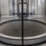Baza unui tunel de vânt vertical, cu podea metalică tip grilaj și pereți circulari din sticlă sau plastic transparent, într-un spațiu interior curat