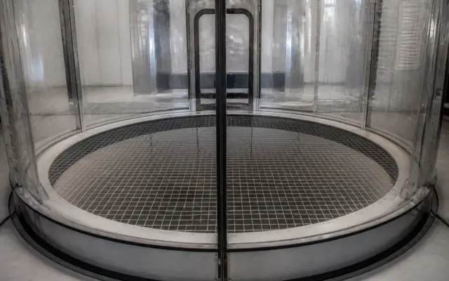 Baza unui tunel de vânt vertical, cu podea metalică tip grilaj și pereți circulari din sticlă sau plastic transparent, într-un spațiu interior curat