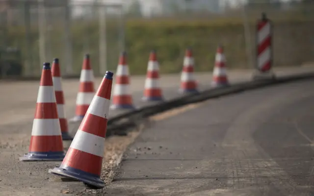 O serie de conuri de trafic alb-roșii așezate în linie de-a lungul marginii unui drum proaspăt asfaltat, delimitând o zonă de lucru