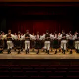 Un grup de dansatori de muzică populară, bărbați și femei, sunt așezați în semicerc pe o scenă de teatru cu podea din lemn și cortină roșie în fundal