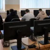 O sală de calculatoare unde mai mulți tineri stau cu spatele la cameră, lucrând la monitoare așezate pe mese lungi de lemn