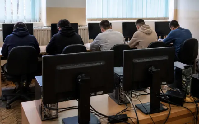 O sală de calculatoare unde mai mulți tineri stau cu spatele la cameră, lucrând la monitoare așezate pe mese lungi de lemn