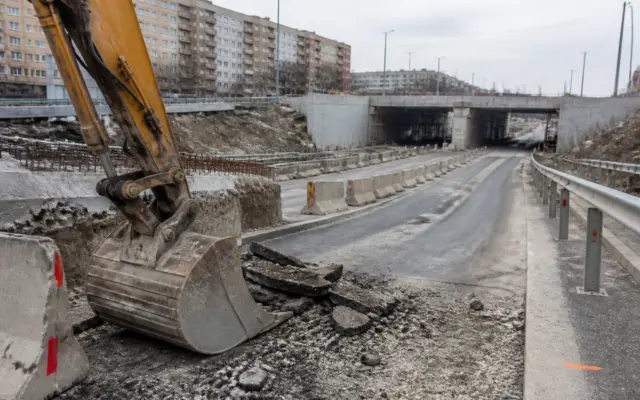 Un braț și o cupă de excavator în prim-planul unui șantier rutier, unde se văd bucăți de asfalt proaspăt excavat