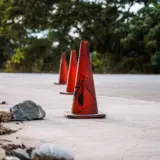 Trei conuri rutiere portocalii sunt așezate în linie pe marginea unei șosele asfaltate, lângă o zonă cu pământ și pietre