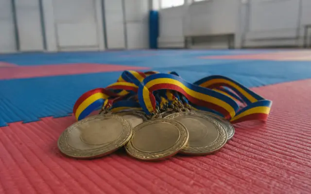 Mai multe medalii aurii și argintii fără inscripții sunt așezate pe o saltea de karate (tatami) în culorile roșu și albastru