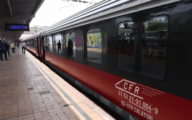 Un vagon de tren CFR Călători, de culoare roșu cu negru, oprit la peronul unei gări