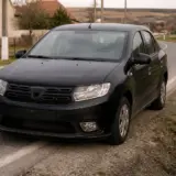 Un autoturism Dacia Logan de culoare neagră este oprit pe marginea unui drum asfaltat, la intrarea sau ieșirea dintr-o localitate rurală