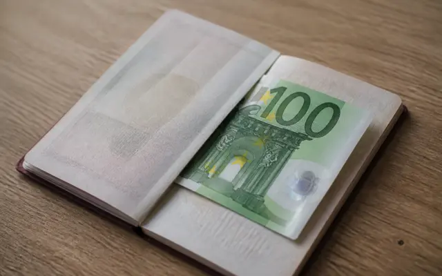 Un pașaport deschis pe o masă de lemn, în interiorul căruia se află o bancnotă de 100 de euro de culoare verde